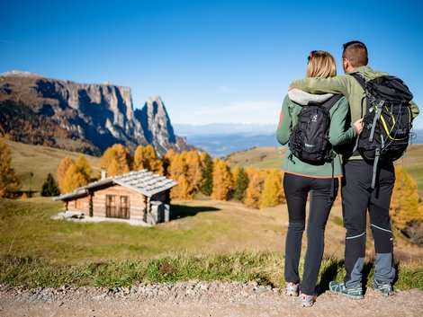Weekly programme of our hotels near Alpe di Siusi Couple with backpacks looking at a cabin and autumn mountains
