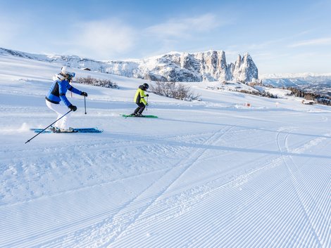 Weekly programme of our hotels near Alpe di Siusi Two skiers on groomed slope with snowy mountains in background