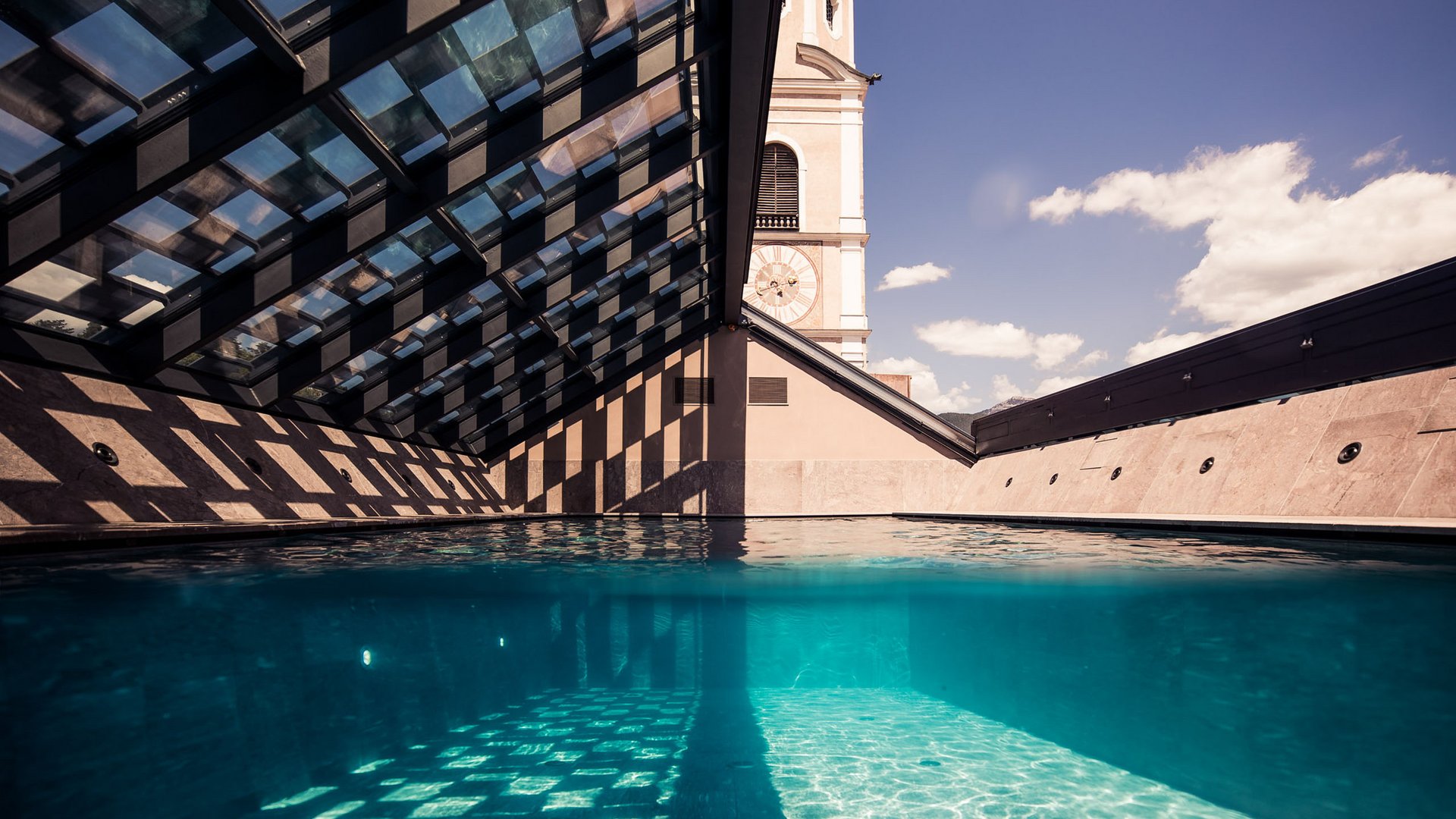 4-Sterne-Hotel Lamm am Fuße der Seiser Alm Schwimmbad mit Glasdach und Turmuhr im Hintergrund bei sonnigem Himmel