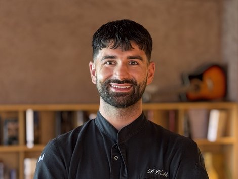 4-Sterne-Hotel Lamm am Fuße der Seiser Alm Lächelnder Mann in Kochjacke mit verschränkten Armen vor Bücherregal