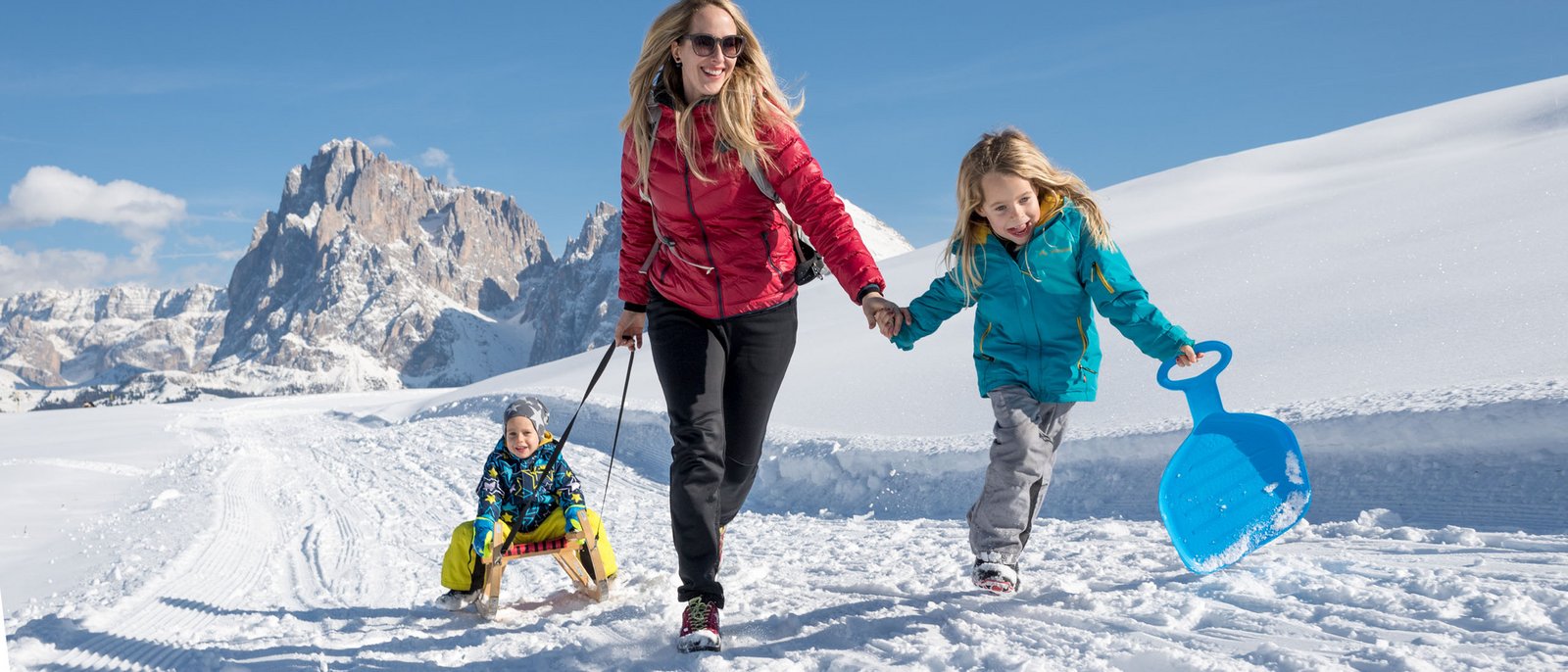 In slittino sull’Alpe di Siusi: il divertimento vi attende! Madre che trascina il bambino sulla slitta in paesaggio montano innevato con figlia e pala