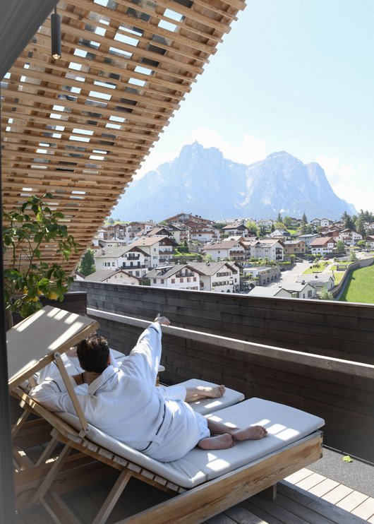4-Sterne-Hotel Lamm am Fuße der Seiser Alm Person entspannt sich auf Balkon mit Bergblick und Häusern im Tal
