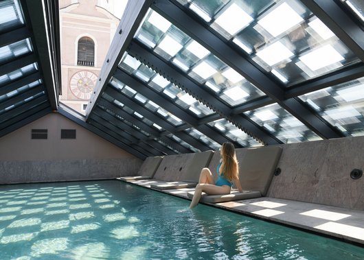 4-Sterne-Hotel Lamm am Fuße der Seiser Alm Frau im blauen Badeanzug sitzt am Rand eines Innenpools unter Glasdach