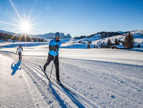 Weekly programme of our hotels near Alpe di Siusi Two people cross-country skiing on snowy landscape under bright sunlight