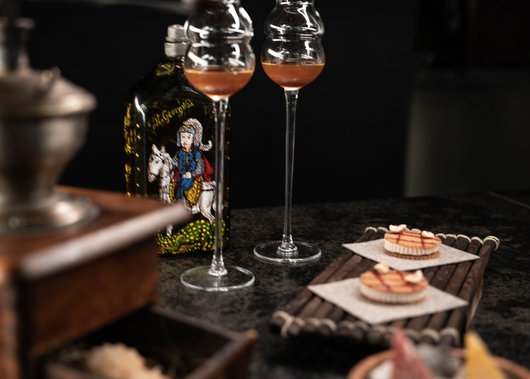 Gourmet restaurant in South Tyrol: Lampl Stube Two liqueur glasses, a bottle with colorful illustration, and small tarts on a tray