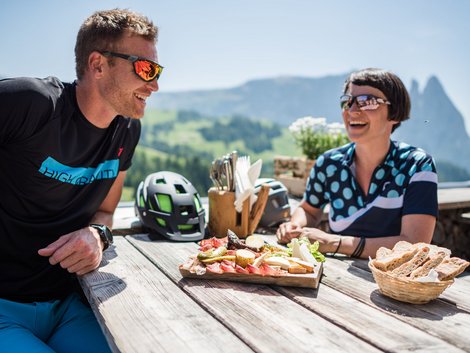 Weekly programme of our hotels near Alpe di Siusi Two cyclists laughing while eating on a sunny terrace with mountain view